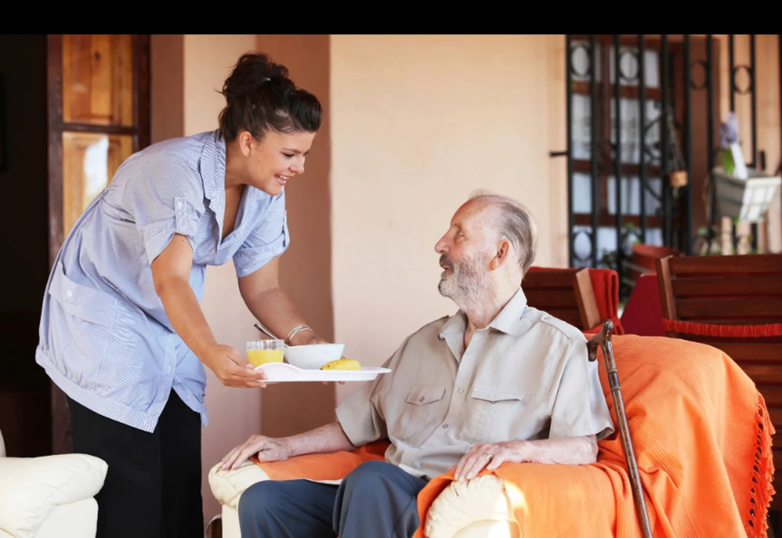 Aidante souriante apportant un plateau-repas à un aîné assis dans son fauteuil à la maison